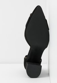 Escarpin en daim noir à talon haut avec un bout pointu, doté d'une semelle en caoutchouc texturée. Le talon a une finition lisse et un arrière légèrement arrondi.