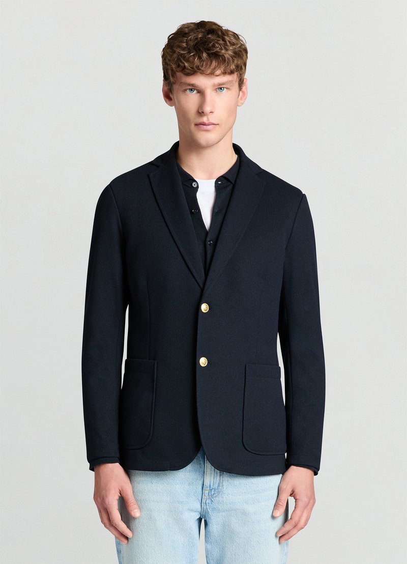 Blazer bleu marine en tissu lisse, avec une coupe ajustée, deux poches avant, et des boutons dorés. Porté sur une chemise à boutons.