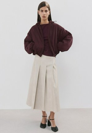 Donna che indossa una giacca imbottita corta color viola scuro, top abbinato, gonna midi plissettata beige e scarpe Mary Jane nere con tacco, in piedi contro un muro bianco.