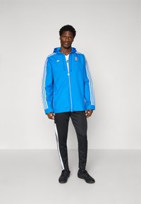 Blå vattentät jacka med huva, vit tre-randig design på ärmarna, logodetalj, kombinerad med svarta byxor med vita ränder.