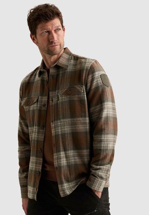 Homme portant une chemise à carreaux marron et grise à boutons avec poches poitrine, regardant sur le côté avec une main dans la poche d’un pantalon noir.