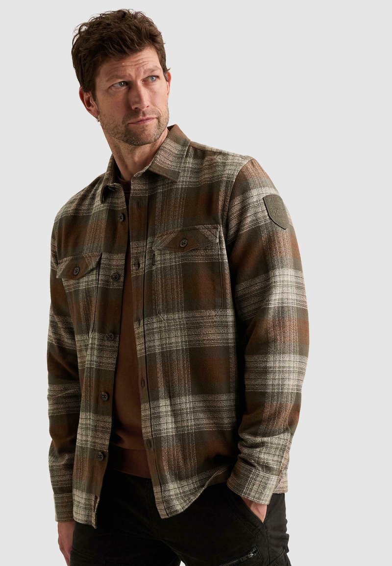 Homme portant une chemise à carreaux marron et grise à boutons avec poches poitrine, regardant sur le côté avec une main dans la poche d’un pantalon noir.