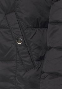 Schwarzer Steppjackenabschnitt mit sichtbaren Nähten, einer diagonalen Naht und einem glänzenden silbernen Druckknopf. Das Material wirkt glatt und isoliert.