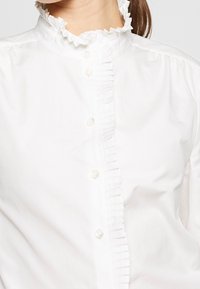 Blouse en coton blanc avec un col froncé, une fermeture à boutons à l'avant avec une patte, et une texture lisse. Des volants soulignent le décolleté.