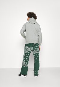 Sudadera con capucha gris y un bolsillo tipo canguro, combinada con pantalones de chándal verdes que presentan un patrón de paisley blanco. Zapatillas negras con detalles blancos.