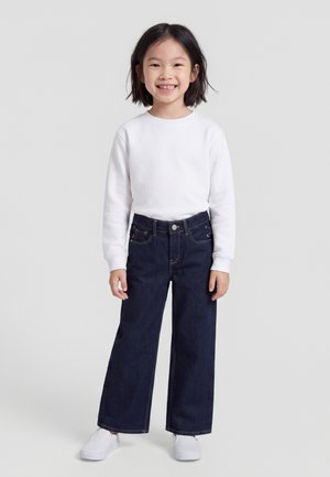 Enfant souriant aux cheveux courts et foncés portant un t-shirt blanc à manches longues, un jean foncé à jambes larges et des baskets blanches, debout sur un fond uni.