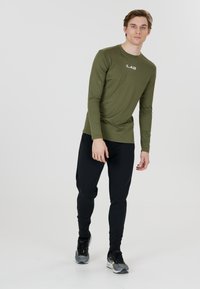Langærmet olivengrøn atletisk top med en tætsiddende pasform og logo. Sammen med sorte tapered joggerbukser og grå/sorte løbesko.