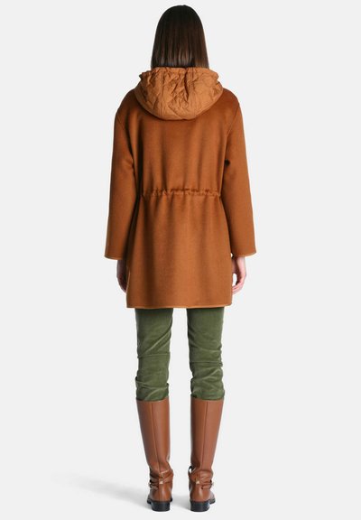 Femme debout de dos, portant un manteau à capuche marron, un pantalon vert olive, et des bottes marron montantes jusqu'au genou sur fond blanc.