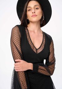 Zwarte jurk met doorschijnende stippenmouwen, V-hals en elastische manchetten. De stof heeft een gladde textuur met een stippenpatroon en een gelaagde rok.