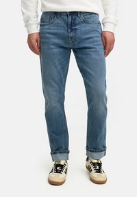 Lyse blå denimjeans med rett passform, synlig falming og rullet opp ermer, kombinert med kremfargede og svarte joggesko.