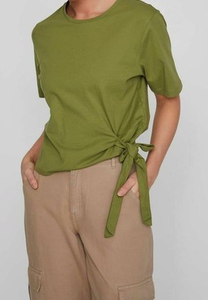 Kobieta ubrana w oliwkowozielony top z krótkim rękawem i wiązaniem po bokach oraz beżowe spodnie cargo, ręce w kieszeniach, neutralne tło.