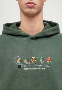 Haki zaļš kapuci ar krāsainiem izšūtiem putniem virs "Abercrombie & Fitch" teksta; mīksta auduma, ar brīvu griezumu un ar šņorēm kapuce.