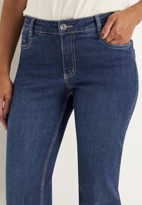 Mörkblå denimjeans med en klassisk rak ben-design, med subtila sömmar och en framskjutande knäppning. Fickor synliga på sidorna.