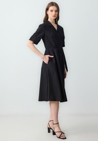 Robe noire à manches courtes, avec une taille ceinturée et des poches latérales. Tissu lisse, longueur en dessous du genou, associée à des talons noirs à lanières.