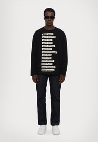 Black sweatshirt with white text phrases on the front, paired with dark jeans and white sneakers. Model wearing black sunglasses.