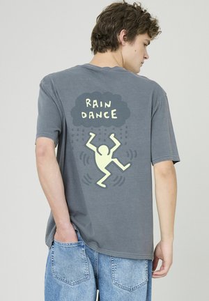 T-shirt en coton gris à manches courtes avec un graphisme jaune au dos portant l'inscription « DANSE DE LA PLUIE » accompagnée d'un danseur abstrait et d'un motif de pluie.