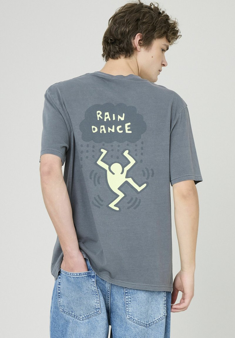 T-shirt en coton gris à manches courtes avec un graphisme jaune au dos portant l'inscription « DANSE DE LA PLUIE » accompagnée d'un danseur abstrait et d'un motif de pluie.