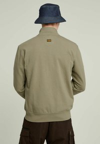 Felpa khaki con collo alto, polsini a costine, texture morbida e un'etichetta quadrata sulla parte alta della schiena; indossata con un cappello a secchiello in denim.