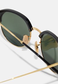 Ochelari de soare cu ramă neagră și lentile verzi, cu accente subțiri din metal auriu și tampoane nazale transparente, având o formă elegantă, rotunjită și un design minimalist.