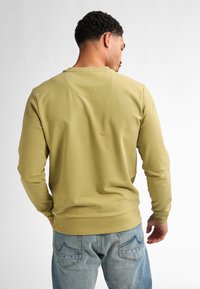 Sudadera de manga larga color verde oliva con cuello redondo y puños acanalados, combinada con unos vaqueros de color azul claro que presentan detalles en los bolsillos traseros.