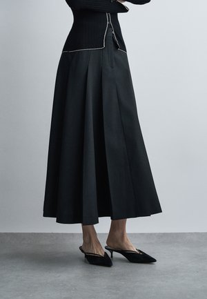 Femme portant une longue jupe plissée noire, des talons slingback noirs à bout pointu et un pull côtelé noir à bordure blanche, debout sur un sol gris.