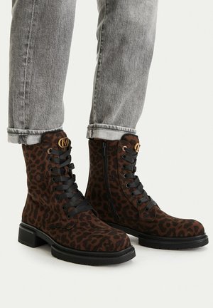 Braune, leopardbedruckte Schnürstiefel mit schwarzen Sohlen, getragen zu hochgekrempelten grauen Jeans auf hellem Hintergrund.