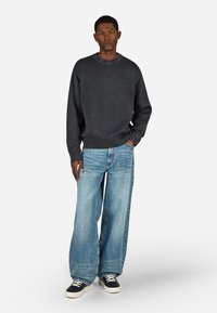 Pelēks adīts džemperis, plata gaismas zila džinsu bikses un melni sporta apavi. Vienkāršs dizains ar brīvu piegriezumu un minimālu detalizāciju.