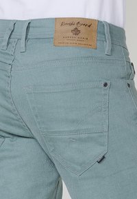 Pantalons en denim bleu-vert clair avec une patch en cuir à la taille, design classique à cinq poches et détails de couture contrastants.