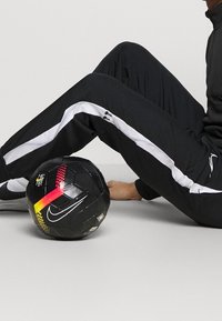Schwarzer Nike Fußball mit bunten Akzenten in Rot und Gelb, der auf einem grauen Boden liegt, nahe einem schwarzen Trainingsanzug mit weißen seitlichen Streifen.