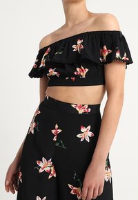 Svart off-shoulder crop top med volangkant och blommönster. Matchande högmidjade byxor har liknande blommönster i rosa och vitt.