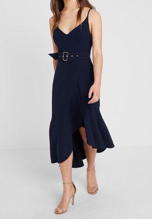 Woman wearing a sleeveless navy blue belted dress with an asymmetrical hem and beige high-heeled sandals, standing on a plain background.