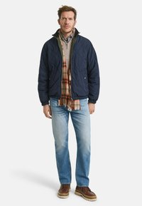 Marineblauwe gewatteerde jas met een ritssluiting, groene voering, gedragen over een beige trui en een geruit overhemd. Lichtblauwe vervaagde jeans en bruine laarzen.