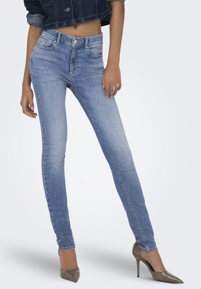 Moteris dėvi šviesiai mėlynas aptemptas džinsines kelnes, tamsų trumpą džinsinį švarką ir rudos spalvos aukštakulnius batelius su smailiu priekiu, stovinti priešais neutralią foną.