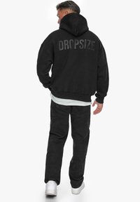 Schwarzer Hoodie mit einem großen, grauen "DROPSIZE"-Druck auf dem Rücken. Kombiniert mit schwarzen Hosen und weißen Sneakers; hat eine Kapuze und gerippte Bündchen.