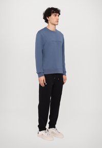 Sudadera azul con bordado del logo en el pecho, combinada con pantalones de chándal negros y zapatillas blancas. La sudadera tiene puños acanalados.