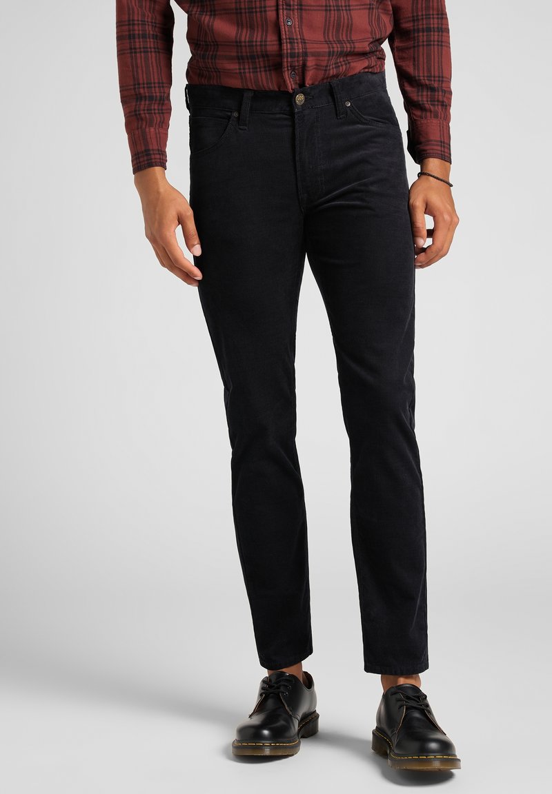 Homme portant un pantalon en velours côtelé noir ajusté, des chaussures en cuir noires et une chemise à carreaux rouge à manches longues, debout devant un fond uni.
