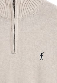 Maglione beige lavorato a maglia con colletto a coste alto, cerniera frontale e un piccolo logo blu ricamato sul lato sinistro del petto. Texture morbida, design casual.