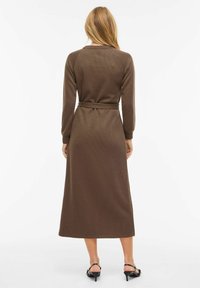 Długa brązowa sukienka z prążkowanego materiału, z paskiem w talii, długimi rękawami i okrągłym dekoltem. Krój dopasowany, o długości midi.