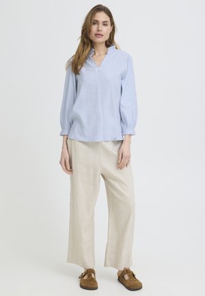 Žena stojaca, oblečená v svetlomodrej blúzke s dlhým rukávom, béžových širokých nohaviciach a hnedých semišových topánkach so sponou na jednoduchom pozadí.