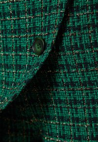 Tissu vert texturé avec des fils noirs et dorés formant un motif à carreaux, avec un bouton vert foncé à quatre trous.