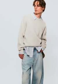 Suéter texturizado color crema superpuesto sobre una camisa de cuadros blancos y negros, combinado con jeans holgados de color azul claro. Anillos notables en los dedos.