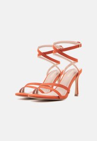 Πέδιλα σε πορτοκαλί σουέντ με ψηλό τακούνι και πολλαπλούς σταυρωτούς λουριά. Διαθέτουν στρογγυλεμένη μύτη και λεπτό τακούνι στιλέτο.