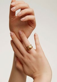 Anillo espiral dorado en una mano con uñas de color rosa claro, sostenido entre dos manos. El fondo es neutro, enfatizando la textura y el diseño del anillo.