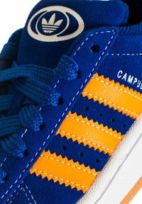 Sneaker blu in suede con strisce in pelle arancione a bordi seghettati, logo rotondo sulla linguetta e lacci texture. Suola in gomma bianca.