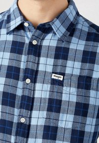 Koszula w kratę z guzikami w odcieniach jasnoniebieskim i granatowym, z kieszonką na piersi i małą białą metką "Wrangler". Miękka tekstura.