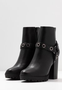 Botas de tobillo de cuero negro con un alto tacón de bloque, cremallera lateral y una correa con acentos metálicos circulares. Suela de goma texturizada.