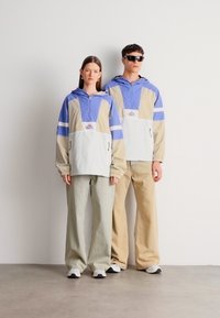 Két modell visel kék, bézs és fehér színblokkos dzsekiket cipzáras zsebekkel, valamint bő szabású, bézs és szürke nadrágokat.