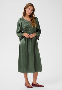 Satin green dress with a smocked bodice, long sleeves, and a V-neck. Paired with burgundy flat shoes. Smooth texture, midi length.