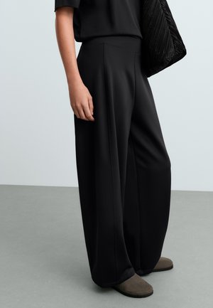 Personne portant un pantalon large noir, un haut noir, tenant un sac matelassé noir, debout sur un sol gris avec un fond gris.