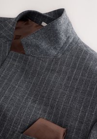 Veste de costume grise à fines rayures avec un col montant et un mouchoir de poche marron, affichée sur un fond clair.